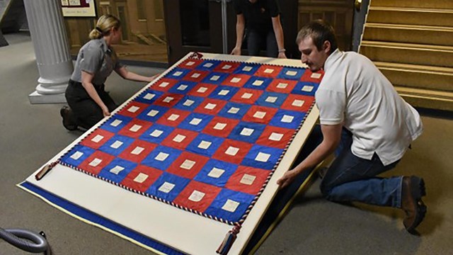 NPS staff bend to carefully move a red and blue quilt laid flat on a large board. 