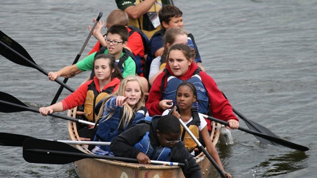 Young kids paddle an extra large canoe on the calm waters of a river.