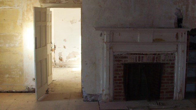 Sun shines through a window on rough cream colored walls. A fireplace is in the shadow. 