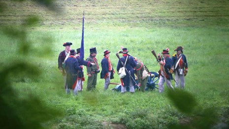 Group of reenactors in blue coats, white breeches & tricorn hats in grassy field by cannon