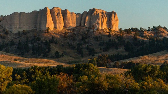 Sandstone buttes rise above grassy field, bathed in orange sunset