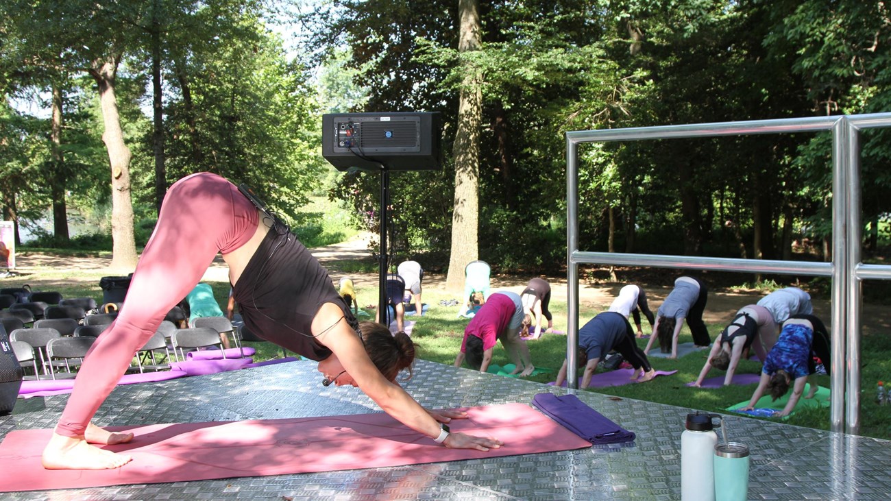 A yoga instructor leads a yoga class in a downward dog pose from an elevated stage