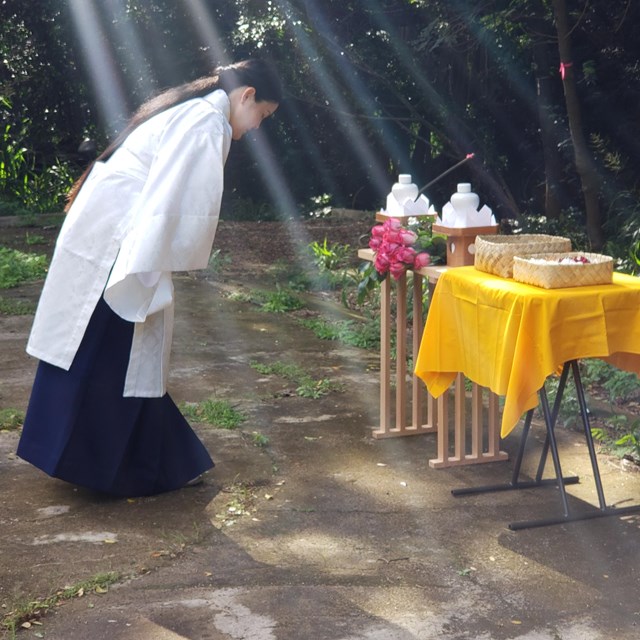 Person outdoors bowing at a small table with various ceremony objects on it 