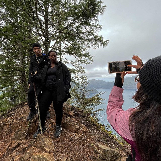 Visitor taking a picture of two hikers next to a tree and lake 