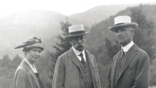 Black and white image of three people in 20th century European style clothes posing on a mowed field