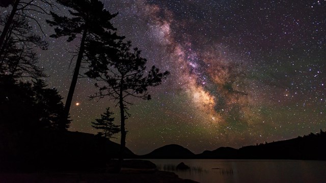 Milky way over a lake with silhouetted trees and mountains