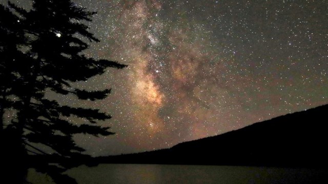 The milky way and other stars over a dark mountain, pond, tree