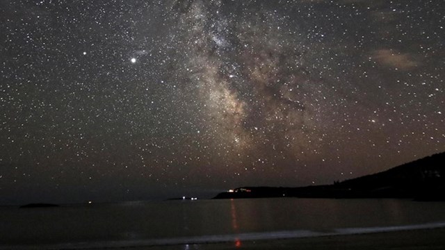 Stars shine over a beach with a distant light on a peninsula 