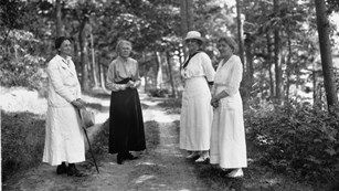 A black and white image of four women in long dresses standing on a forest path.