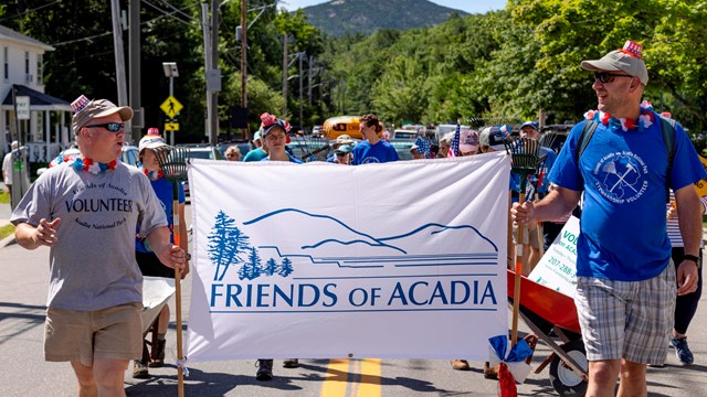 multiple people walking down street in parade carrying Friends of Acadia banner