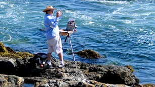 Painter with small easel uses a smartphone to snap a photo up the coastline