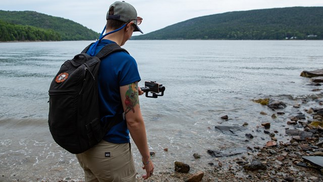 A person holds a handheld phone stabilizer to record footage of a lake