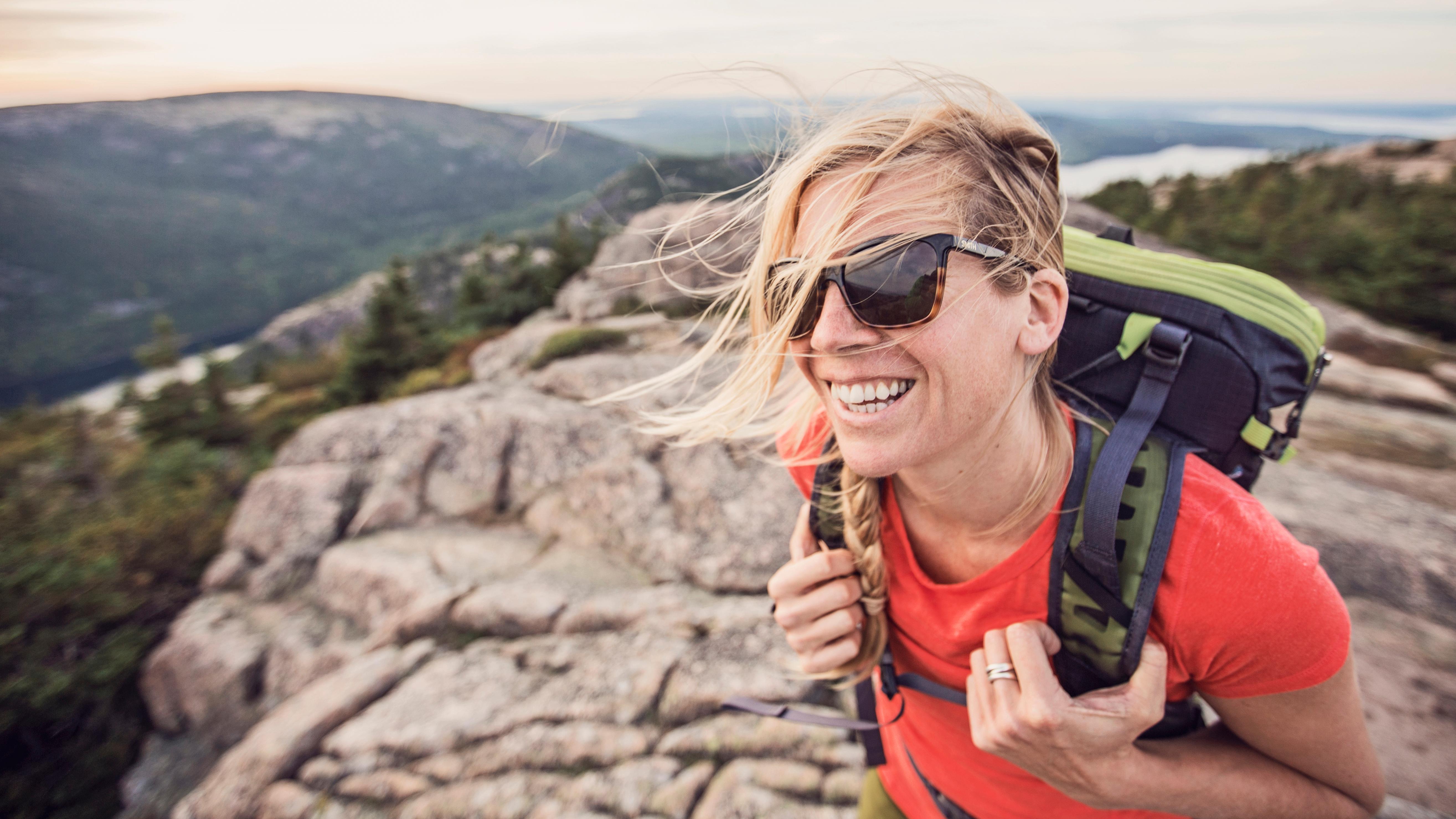 Woman wearing backpack and sunglasses smiles on mountain summit with wind in blowing across her face