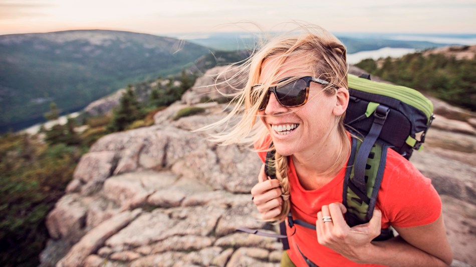 Woman wearing backpack and sunglasses smiles on mountain summit with wind in blowing across her face