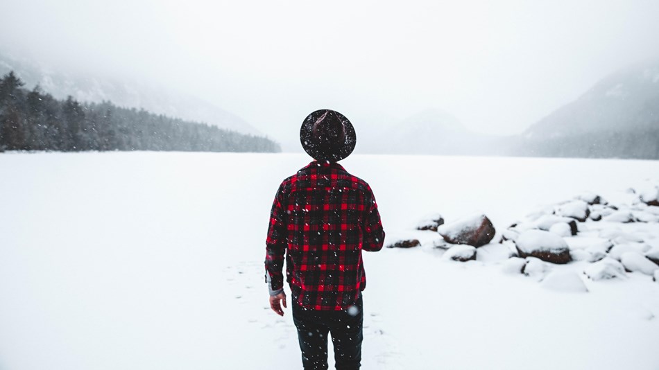 Man in hat and plaid jacket looks across frozen pond as snow falls