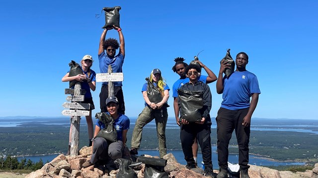 Teenagers and young adults stand on a rocky summit carrying bags.