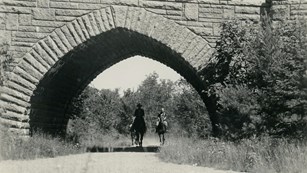 Two people riding horseback under stone bridge