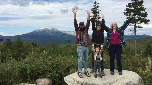 Four people stand with arms outstretched in foreground with mountains in background.