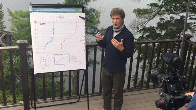 A person films a presenter standing by a flip chart on a wooden deck