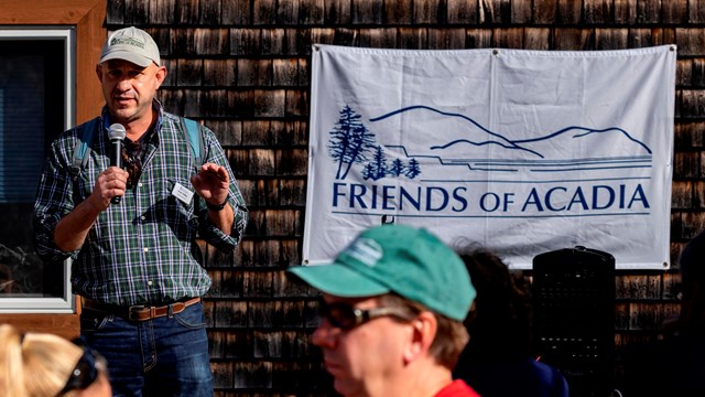 Man in ball cap speaks into a microphone with a Friends of Acadia banner over his shoulder
