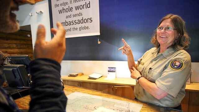 A volunteer standing behind a desk signs to a person with only their arms in frame