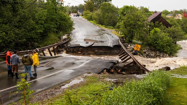 A group of people stand in front of a highway bridge destroyed by a flood.