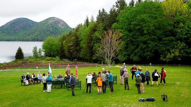 People standing in front of a podium on a lawn with a lake in the background