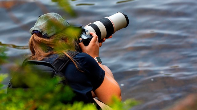 Person holding a camera with large telephoto lens by a lake