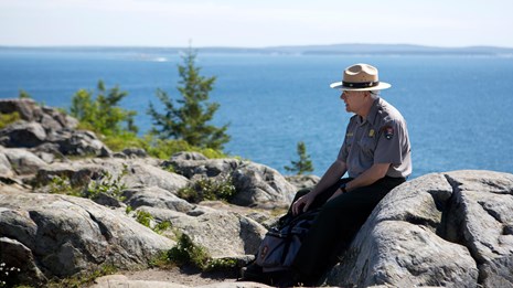 A park ranger sits on a rock along a trail near the coast