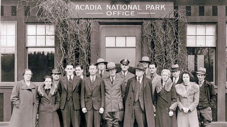 Men & women stand in front of a building, some in uniform; B&W photo