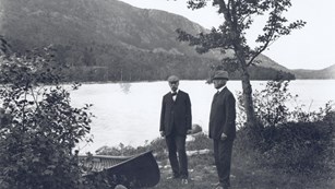 B&W photo of two men with pond and treed hillside behind them