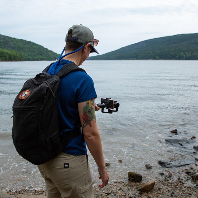 A person holds a handheld phone stabilizer to record footage of a lake