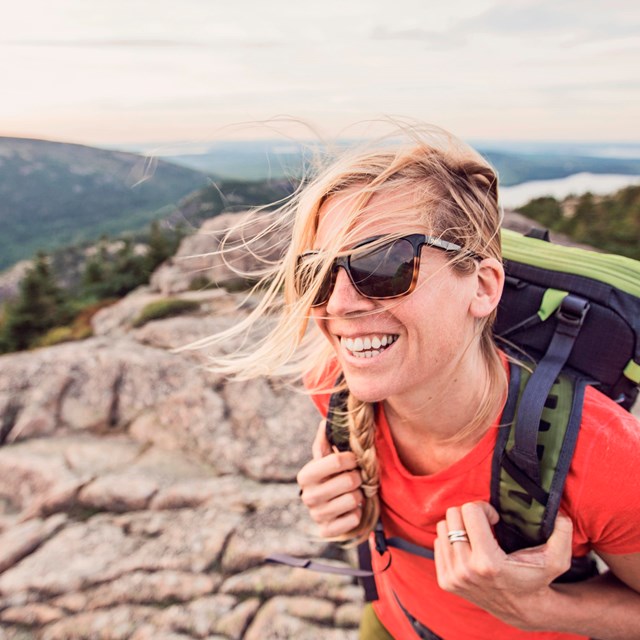 Woman wearing backpack and sunglasses smiles on mountain summit with wind in blowing across her face