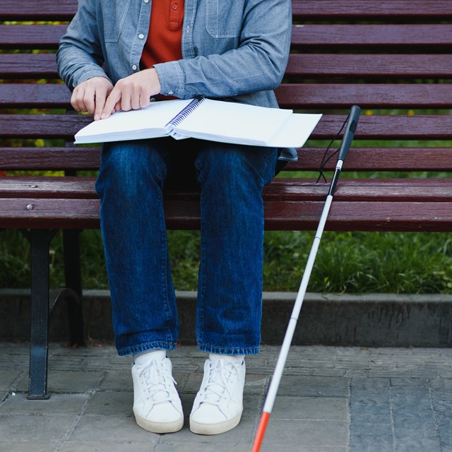 Person seated on a park bench with a red-tip cane uses their hands to read a braille book
