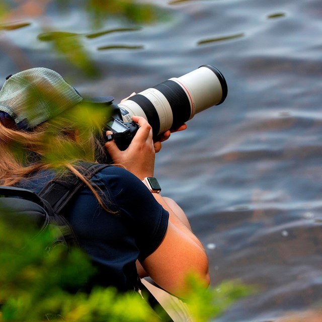 Person holding a camera with large telephoto lens by a lake