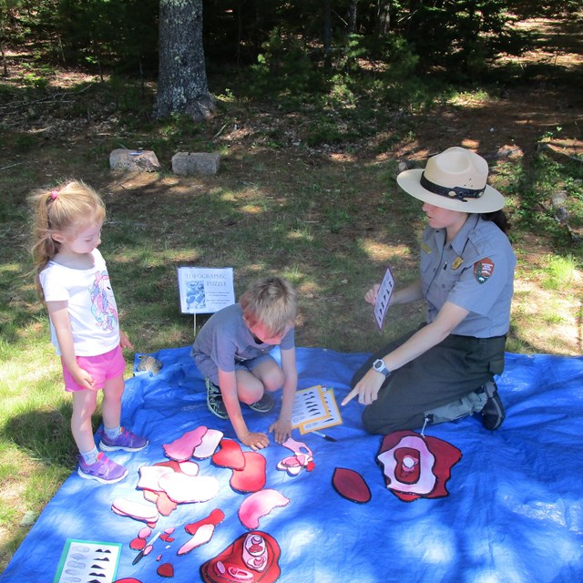 2 children and a ranger sit on a blue mat playing with colored cardboard. 