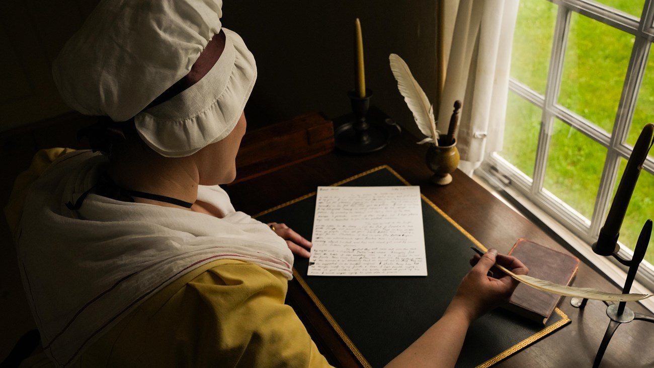 A woman wearing a dress and cap in the 1700s style writes a letter with a quill pen