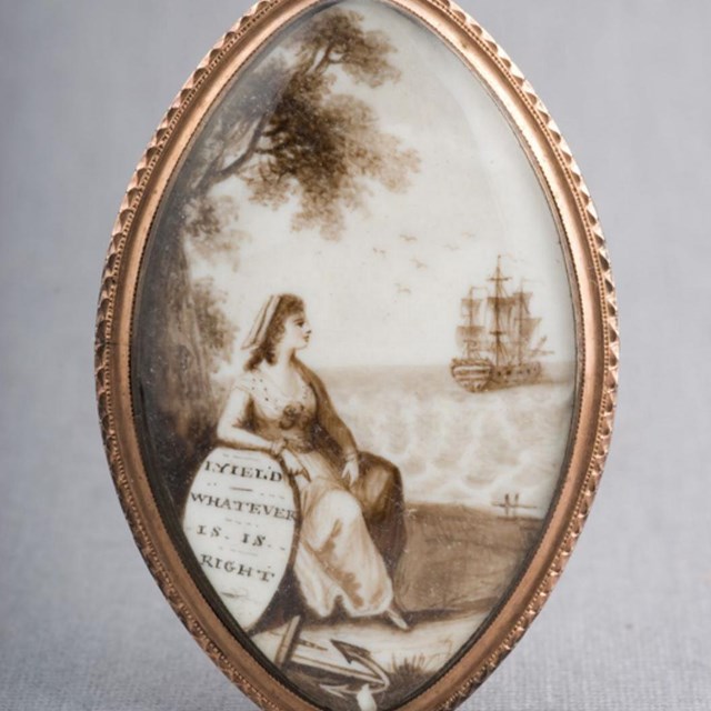 Front face of a gold locket, depicting a woman sitting on a beach leaning on a shield.