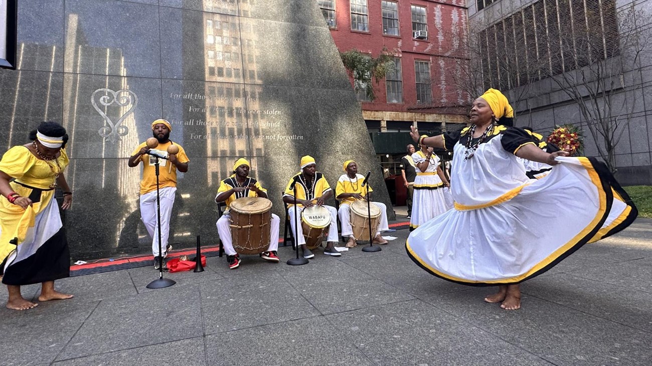 Peformers in yellow, white, and black garb dance in front of the monument. 