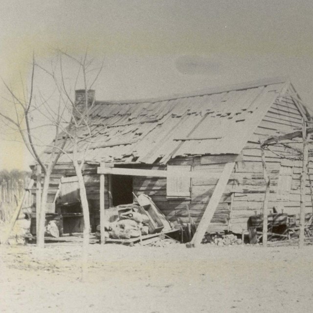 B&W photo of dilapidated cabin; women works outside under a shelter