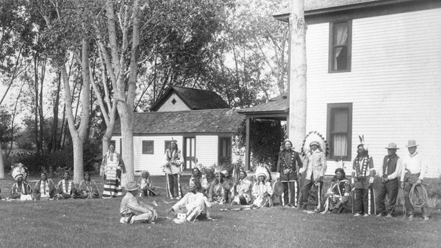 Black and white photo of a gathering of American Indians and Cook at the Agate Springs ranch house.