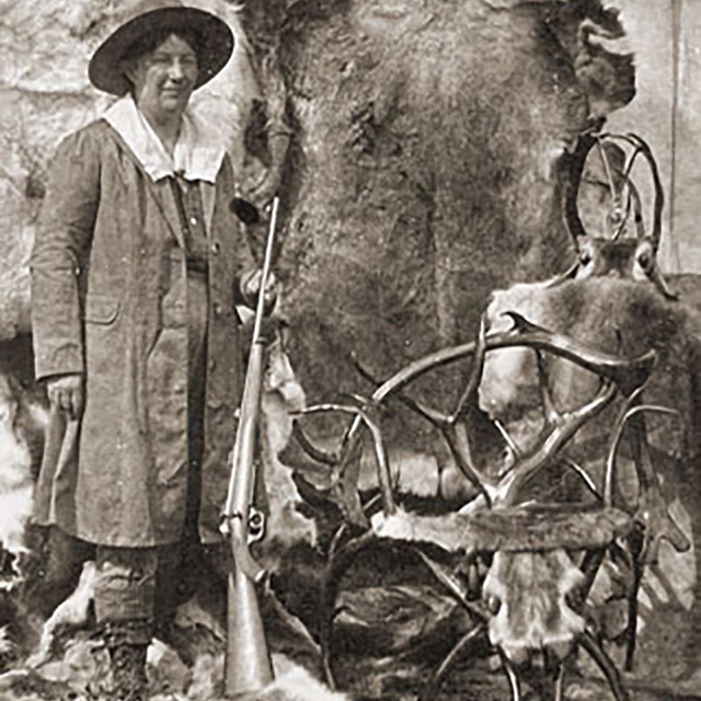 A woman poses with chairs made from antlers of caribou. She is holding a rifle.