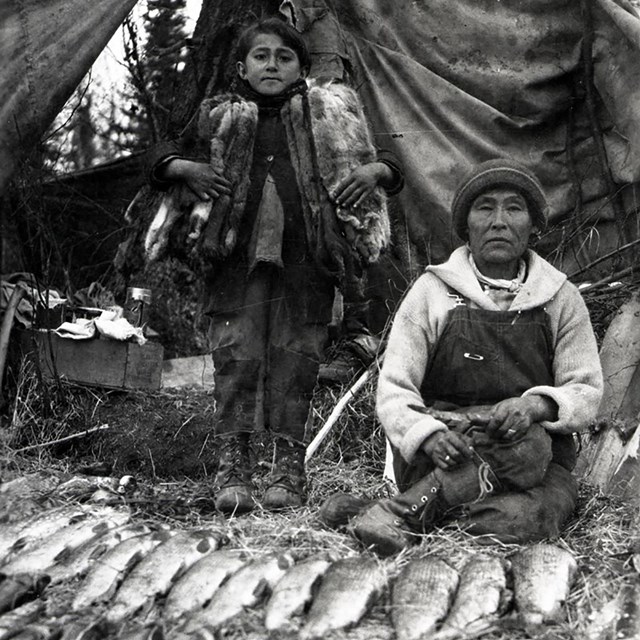 A woman sits on ground with fish in front of her. A young girl stands behind her.