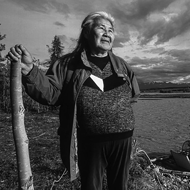 A woman stands with a staff overlooking a river. A fishwheel is in the background.