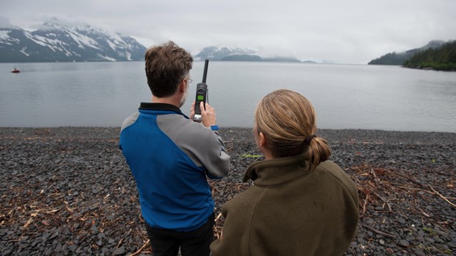 two people use a satellite phone standing on a rocky beach
