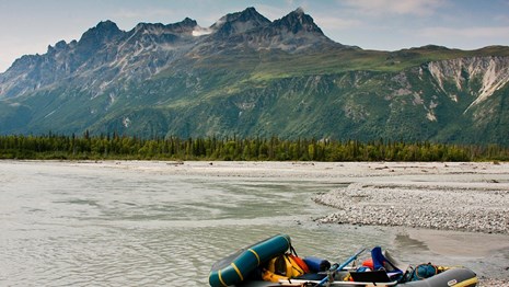 a raft packed with gear sits at the edge of a slow-moving river, with mountains in the background