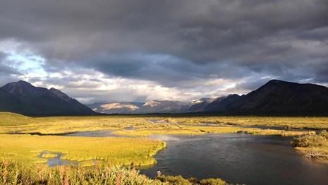 An image of a landscape with grasslands and water. Mountains in background, cloudy day.
