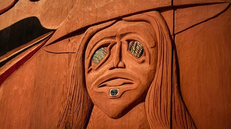 Close up of a carving in wood that shows a face. There are decorative pieces embedded in face.