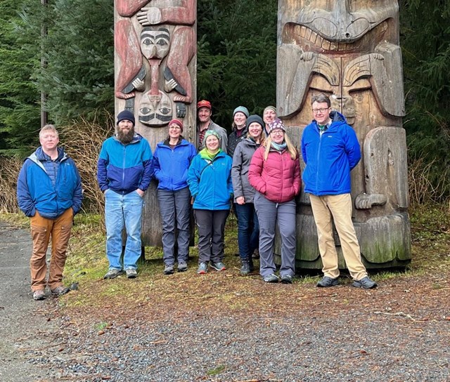 Group of people smiling in front of Alaska Native totem poles