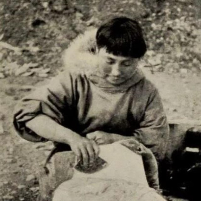 A woman with short hair uses a broad, sharp tool to scrape an animal hide.